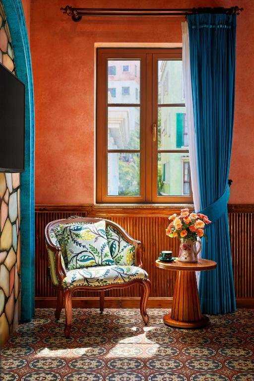 A leaf-print armchair and a small round table with a jug of roses beside a tall window, framed by a blue curtain
