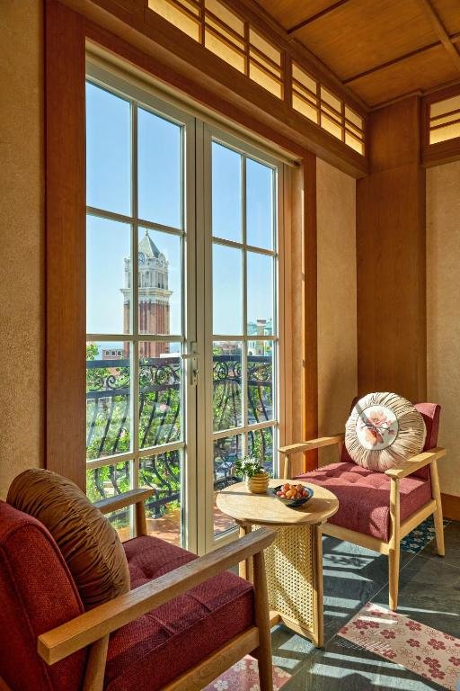 French doors onto a wrought-iron balcony framing the Sunset Town clock tower, with two armchairs and a round table in the sun