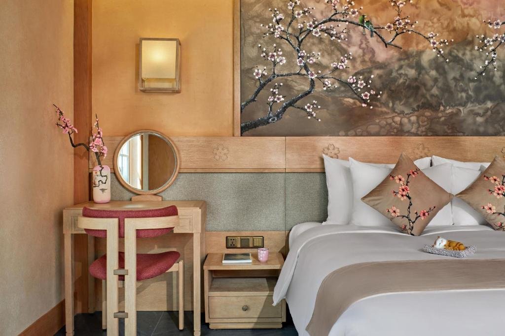 A dressing table with a round mirror and a vase of blossom beside the bed, under a painted blossom panel