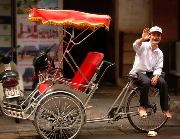 A cyclo driver sitting on his saddle, waving, beside the empty red passenger seat