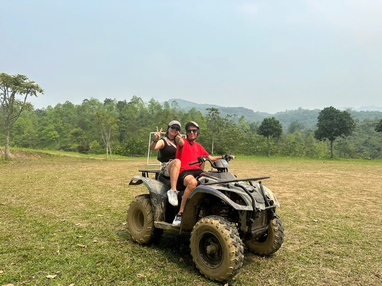Two guests on a quad in an open field in Hoa Binh, forested hills behind them
